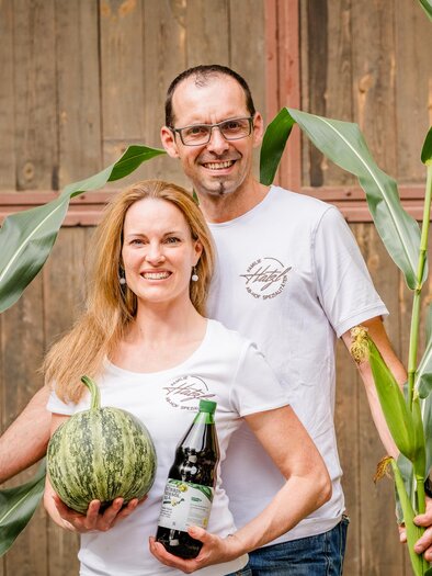 Ein Paar steht zwischen hohen Pflanzen und hält eine Melone sowie eine Flasche in der Hand. Der Hintergrund zeigt eine Holzwand, die eine ländliche Atmosphäre vermittelt. | © Region Graz - Mias Photoart