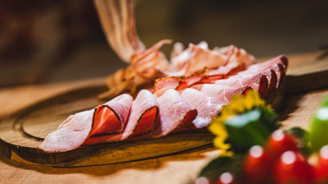 Ein schön angerichtetes Brett mit aufgeschnittenem Schinken. Neben dem Schinken liegen frische Tomaten und Kräuter. | © Region Graz - Mias Photoart