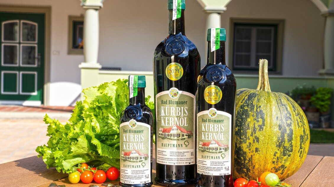 Drei Flaschen Kürbiskernöl stehen auf einem Tisch, umgeben von frischem Salat, Tomaten und einem Kürbis. Im Hintergrund ist ein grünes Gebäude zu sehen. | © Hofladen Hauptmann