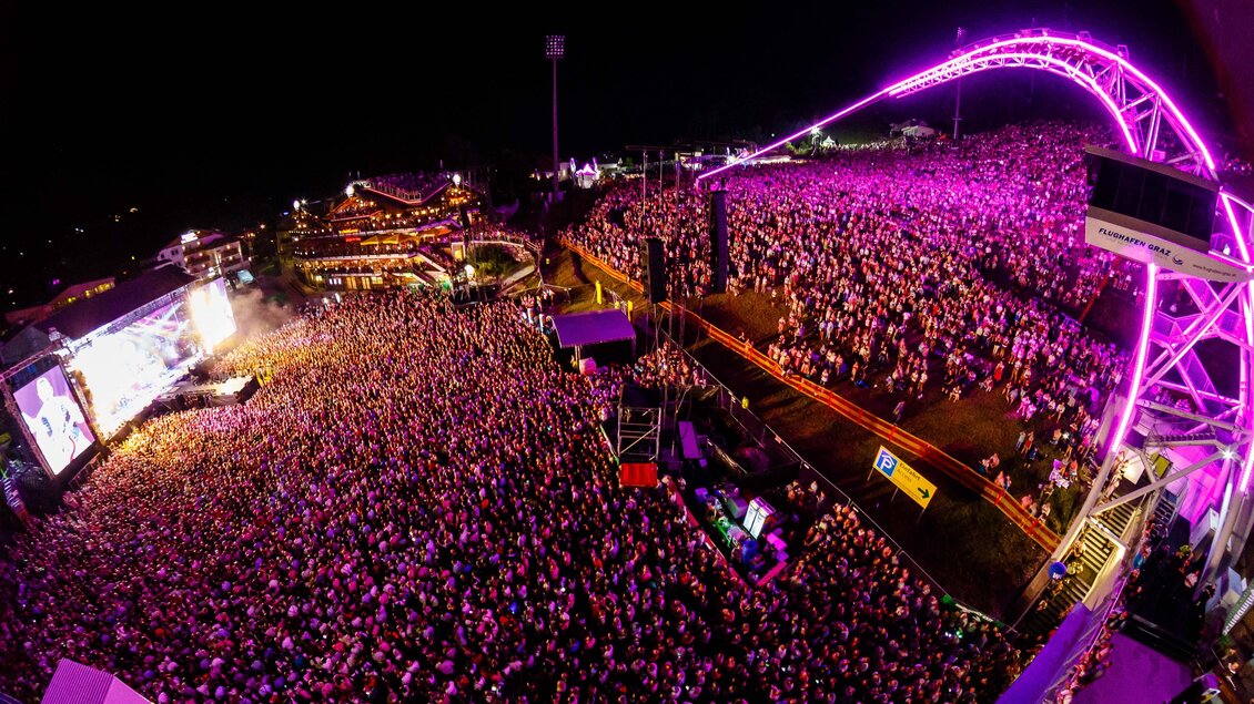 Ein großer Musikfestival mit einer riesigen Menschenmenge unter einer beeindruckenden Lichtshow. Die Bühne ist beleuchtet und die Atmosphäre ist lebhaft und energiegeladen. | © Pressefoto Scharinger