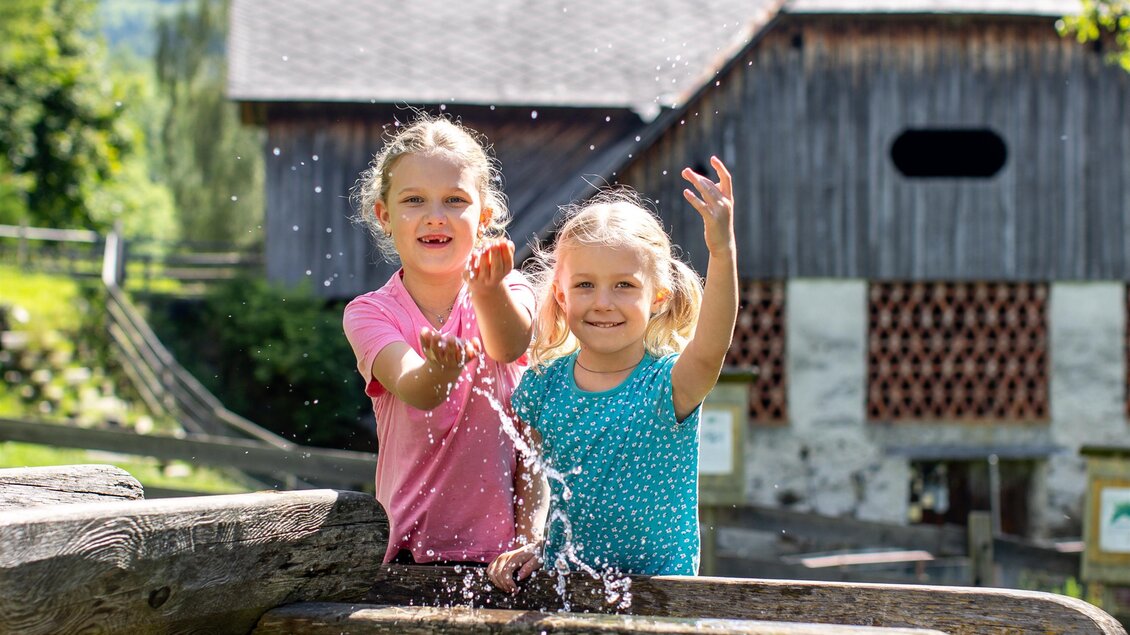 Zwei fröhliche Mädchen spielen am Wasser und spritzen sich nass. Im Hintergrund ist ein Holzgebäude und eine grüne Landschaft zu sehen. | © TVB Murau