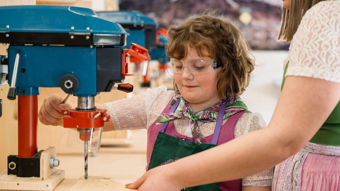 Ein Mädchen arbeitet mit einer Bohrmaschine, während eine Erwachsene ihr hilft. Beide tragen Schutzbrillen und sind in einem handwerklichen Umfeld. | © Narzissendorf Zloam