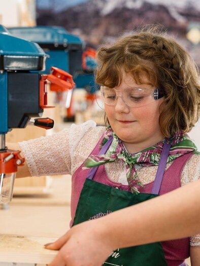 A girl is working with a drill while an adult is helping her. Both are wearing safety goggles and are in a craft environment. | © Narzissendorf Zloam