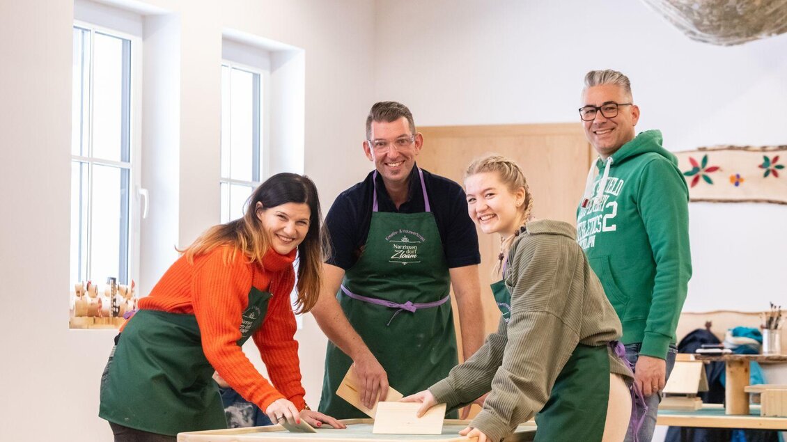 Eine Gruppe von vier Personen arbeitet gemeinsam an einem Holzbearbeitungsprojekt. Sie lächeln und tragen Schürzen in einem hellen Werkraum. | © Narzissendorf Zloam
