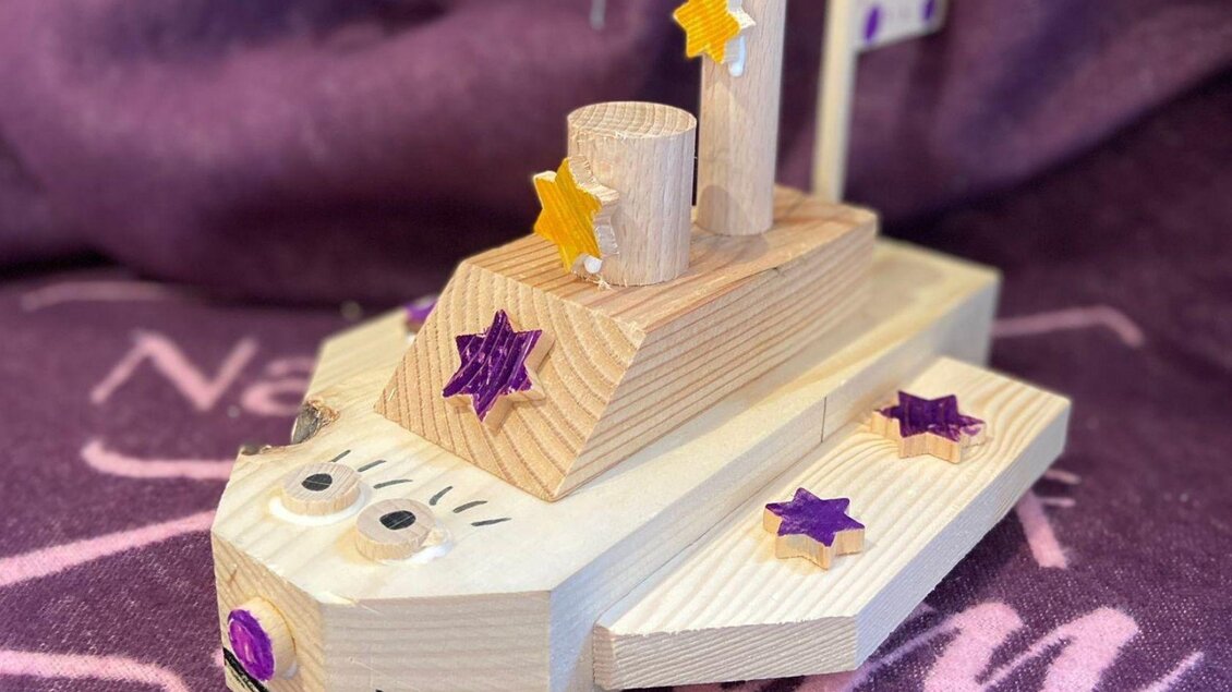 Ein kreatives Holzspielzeug in Form eines Bootes, verziert mit bunten Sternen. Das Spielzeug hat ein fröhliches Gesicht und besteht aus mehreren Holzstücken. | © Narzissendorf Zloam