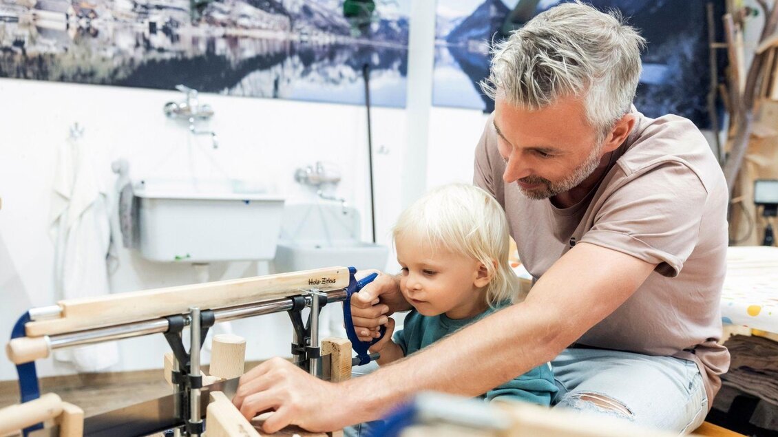 Ein Vater arbeitet mit seinem kleinen Kind in einer Werkstatt. Sie benutzen gemeinsam Holzwerkzeuge und lernen handwerkliche Fähigkeiten. | © www.zloam.at