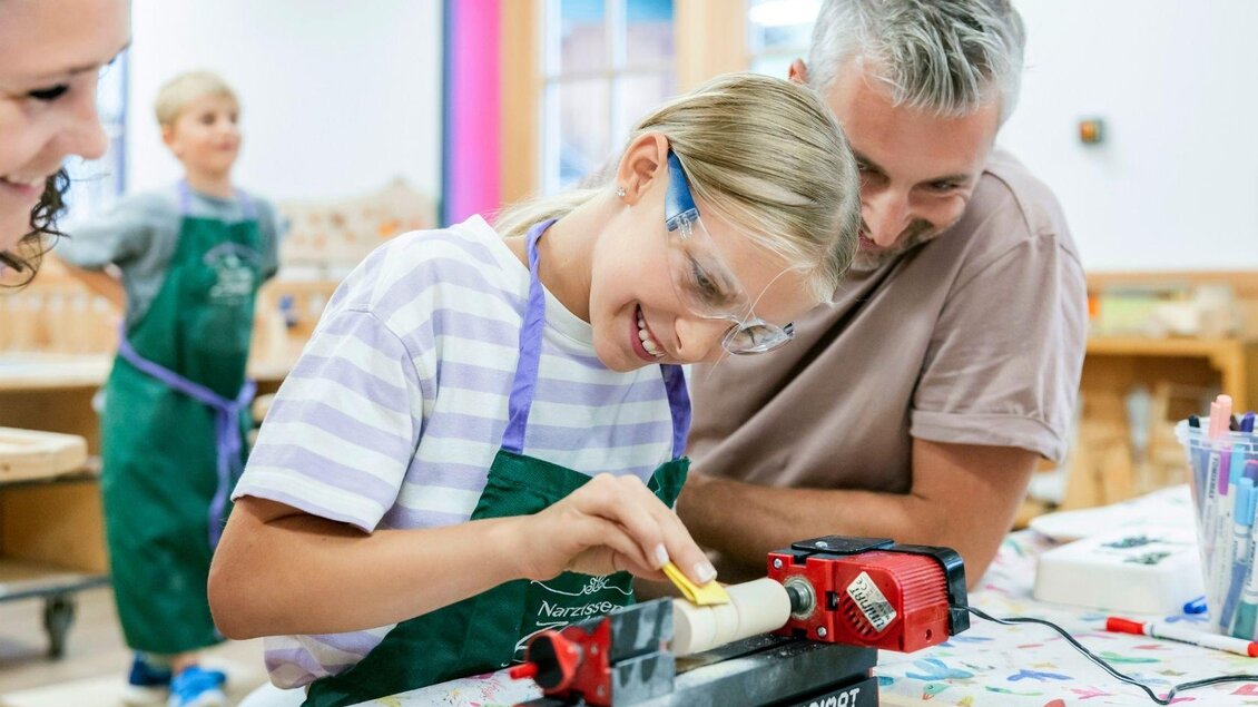 Ein fröhliches Kind arbeitet mit einem Lehrer an einem Holzprojekt. Beide tragen Schutzbrillen und konzentrieren sich auf ihre kreative Aufgabe. | © www.zloam.at