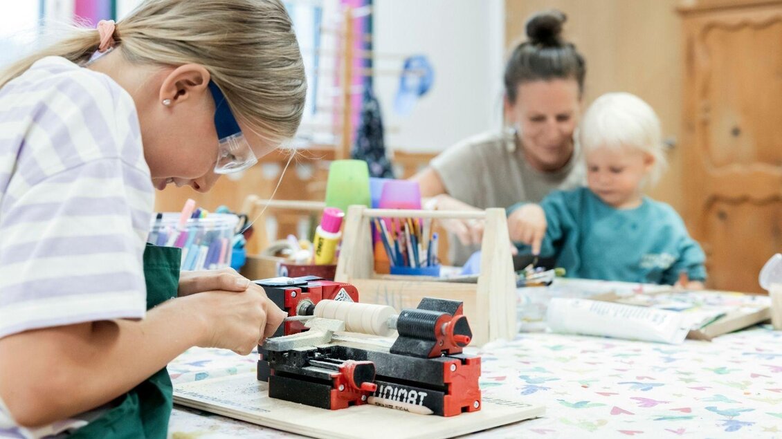 Ein Kind arbeitet an einem Holzprojekt mit einer Werkzeugmaschine. Im Hintergrund sind eine weitere Person und ein kleines Kind zu sehen, die ebenfalls beschäftigt sind. | © www.zloam.at