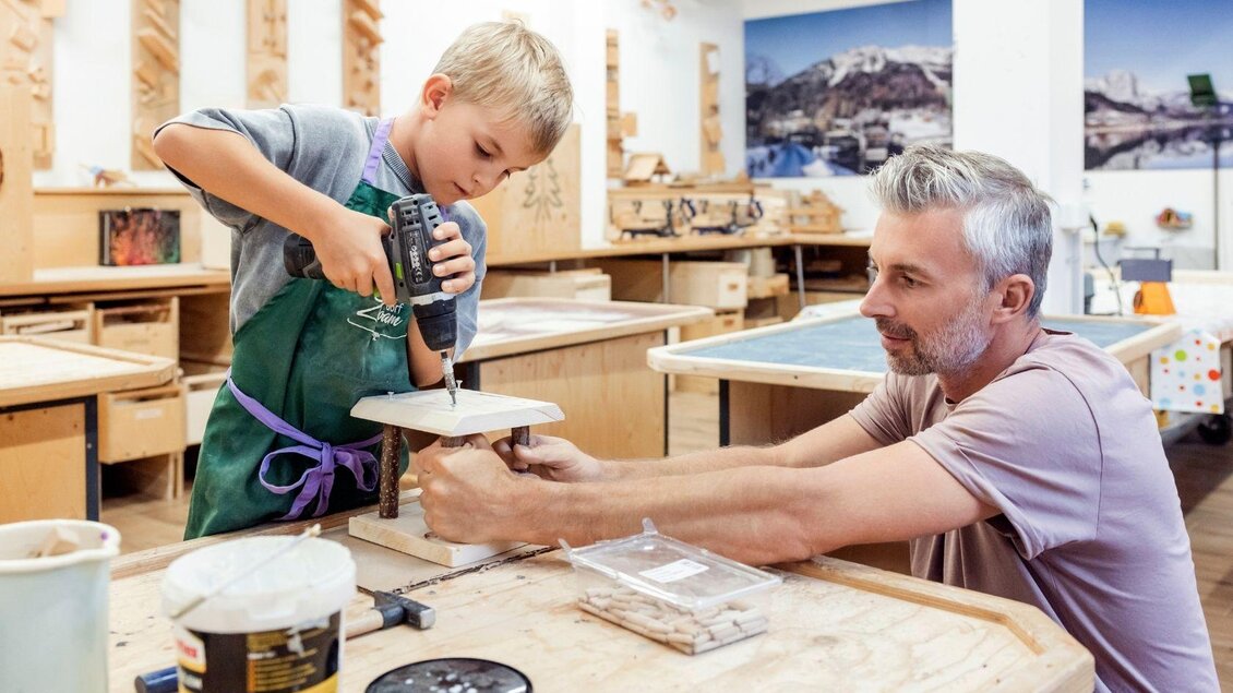 Ein Vater zeigt seinem Sohn, wie man mit einem Bohrer arbeitet. Sie sind in einer Holzwerkstatt umgeben von Werkbänken und Werkzeugen. | © www.zloam.at