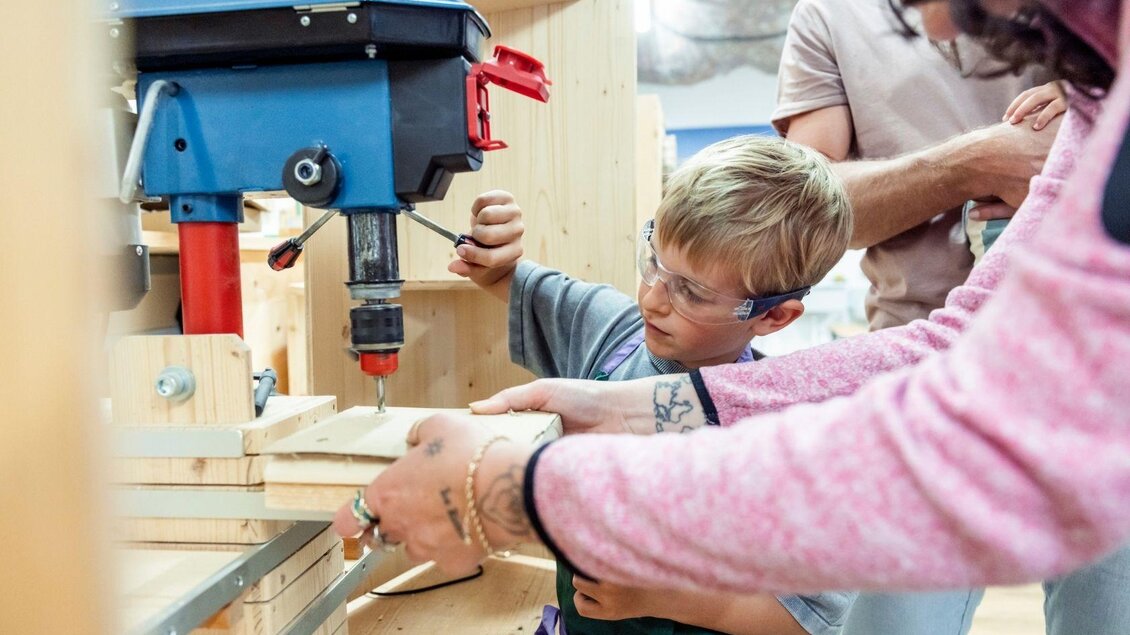 Ein Junge arbeitet mit einem Bohrer in einer Holzwerkstatt. Er wird von einem Erwachsenen unterstützt, während sie gemeinsam an einem Projekt arbeiten. | © www.zloam.at