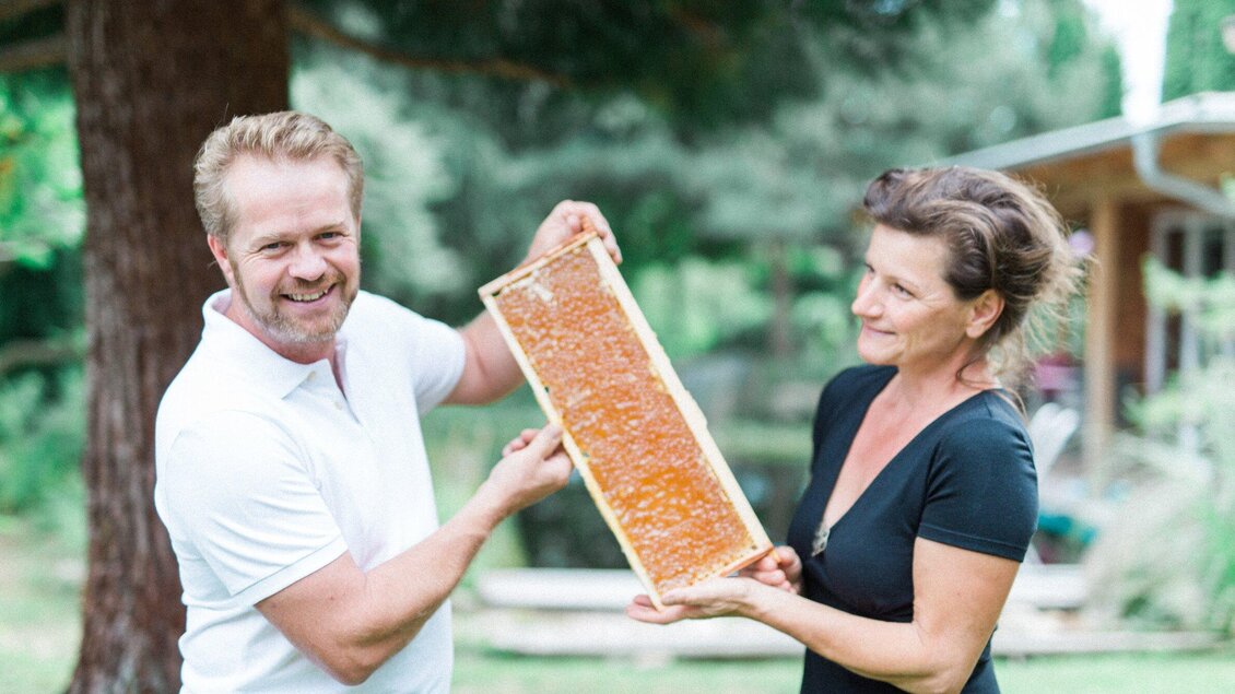 Ein Mann und eine Frau halten gemeinsam einen Rahmen mit frischem Honig. Im Hintergrund sind Bäume und ein Gebäude zu sehen. | © Honiggenuss Manninger