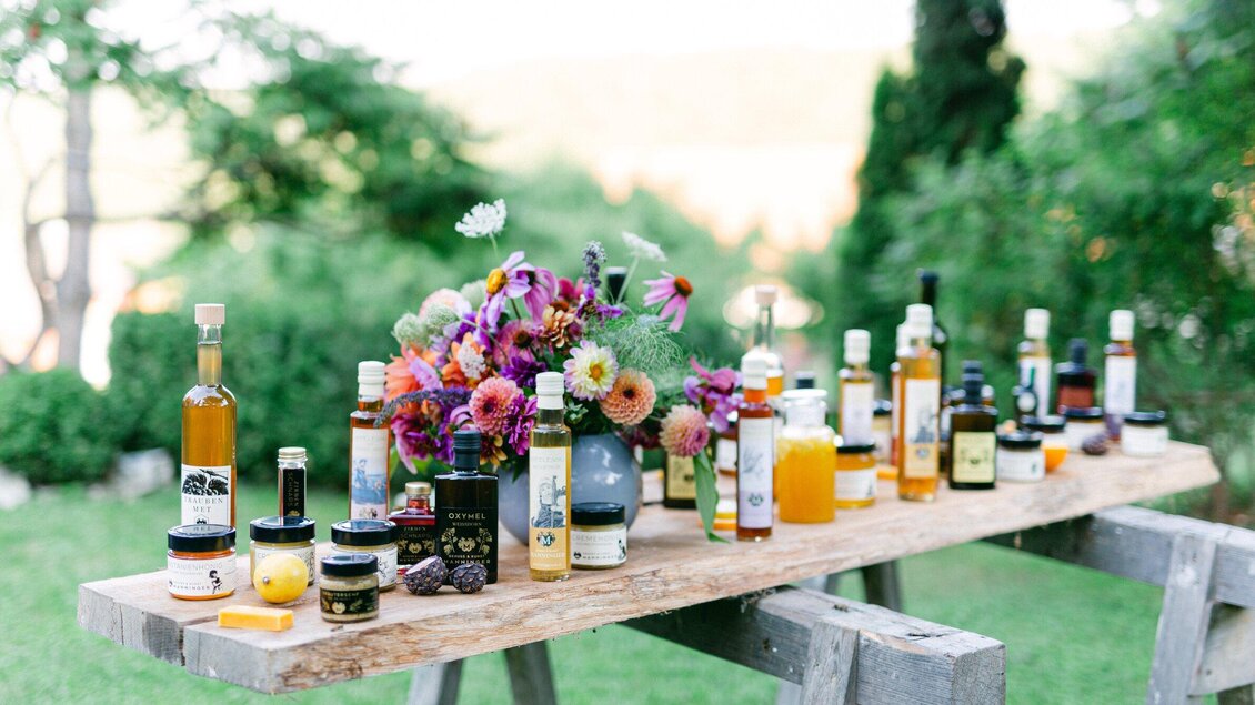 Ein rustikaler Holztisch im Freien, der mit verschiedenen Flaschen von Ölen und Dips dekoriert ist. Bunte Blumenarrangements bringen frische Farben in die Szene. | © Honiggenuss Manninger