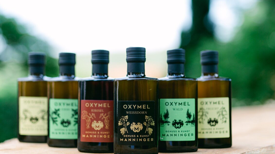 Eine Auswahl an Flaschen mit Oxymel, die verschiedene Geschmacksrichtungen repräsentieren. Die Flaschen stehen auf einem Holzuntergrund mit grüner Natur im Hintergrund. | © Honiggenuss Manninger
