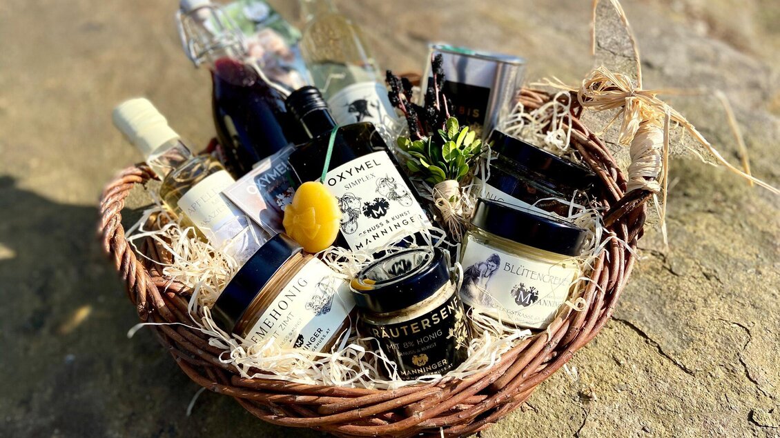 Ein Geschenkkorb mit verschiedenen Gourmetprodukten, einschließlich Gläsern mit Konfitüre, Honig und Flaschen mit Essig. Der Korb ist mit Heu gefüllt und dekoriert mit einer kleinen Zitrone und einem Kräuterauschnitt. | © Honiggenuss Manninger