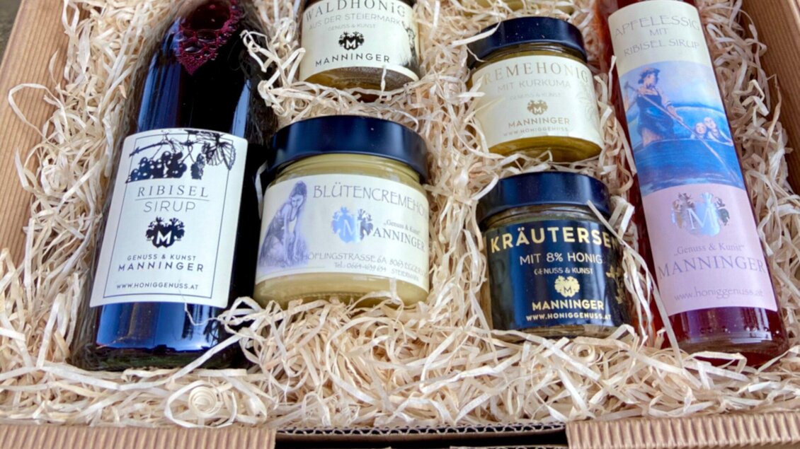 Ein Geschenkkorb mit verschiedenen Produkten, darunter Sirup, Honig und Kräuter. Die Artikel sind sorgfältig in Stroh ausgelegt und in einer schönen Verpackung präsentiert. | © Honiggenuss Manninger