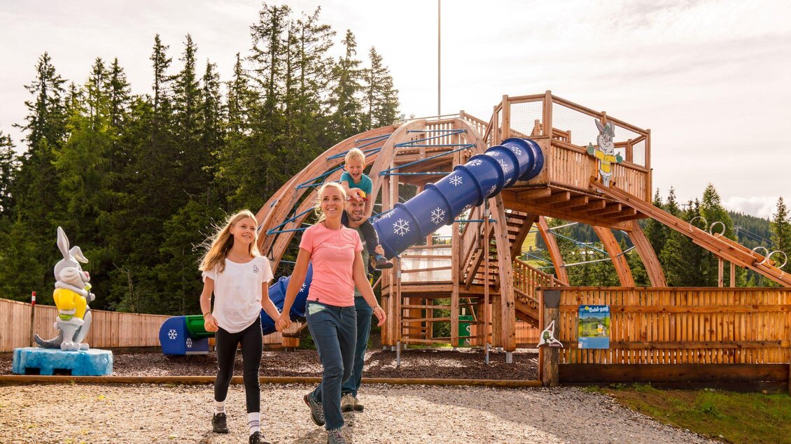 Ein Spielplatz mit einer Rutsche und einem großen Spielturm im Hintergrund. Kinder laufen fröhlich in der Nähe des Spielplatzes und genießen die Zeit im Freien. | © Christine Höflehner