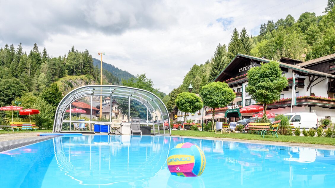 Ein blauer Swimmingpool mit einem bunten Ball im Vordergrund. Im Hintergrund sind Bäume und ein Hotelgebäude mit Sitzbereichen zu sehen. | © Hotel Taferne Swimming Pool