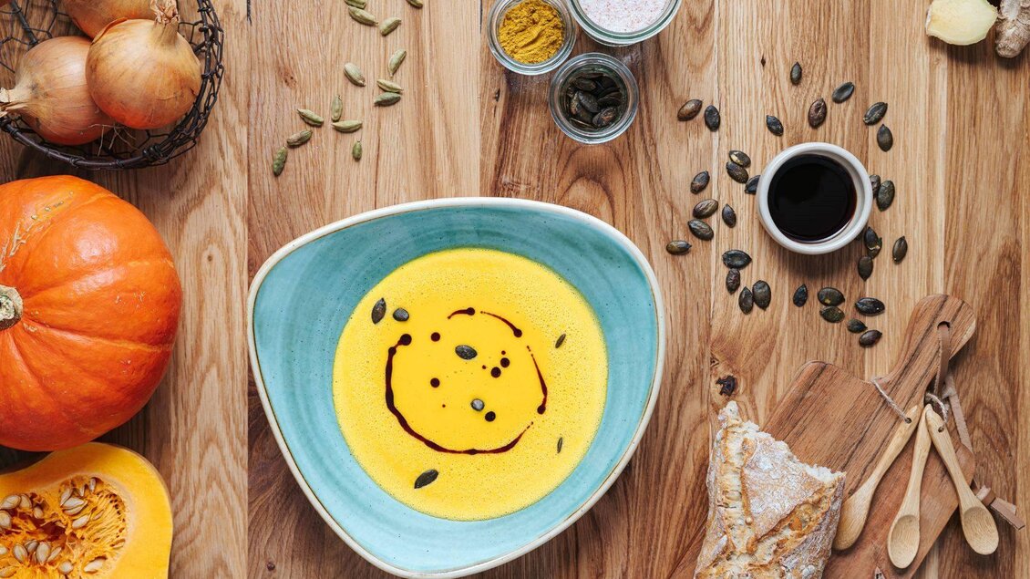 Eine cremige Kürbissuppe in einer blauen Schüssel, umgeben von frischen Zutaten wie Zwiebeln, Kürbis und Gewürzen. Holzbesteck und Brotstücke vervollständigen das rustikale Ambiente. | © Karl Steinegger