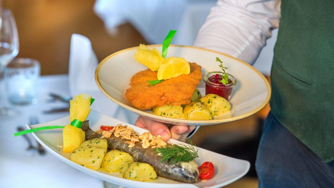 Ein Kellner serviert zwei Teller mit köstlichen Gerichten. Der obere Teller enthält gebratenen Fisch mit Beilagen, der untere Teller zeigt ein panierter Fisch mit Kartoffeln und einer Sauce. | © Karl Steinegger