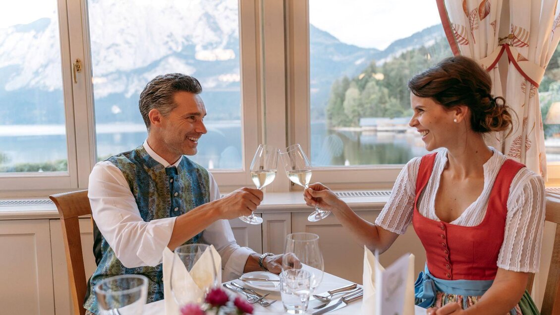 Ein elegantes Dinner-Paar stößt mit Gläsern an. Im Hintergrund ist eine malerische Berglandschaft zu sehen. | © Karl Steinegger