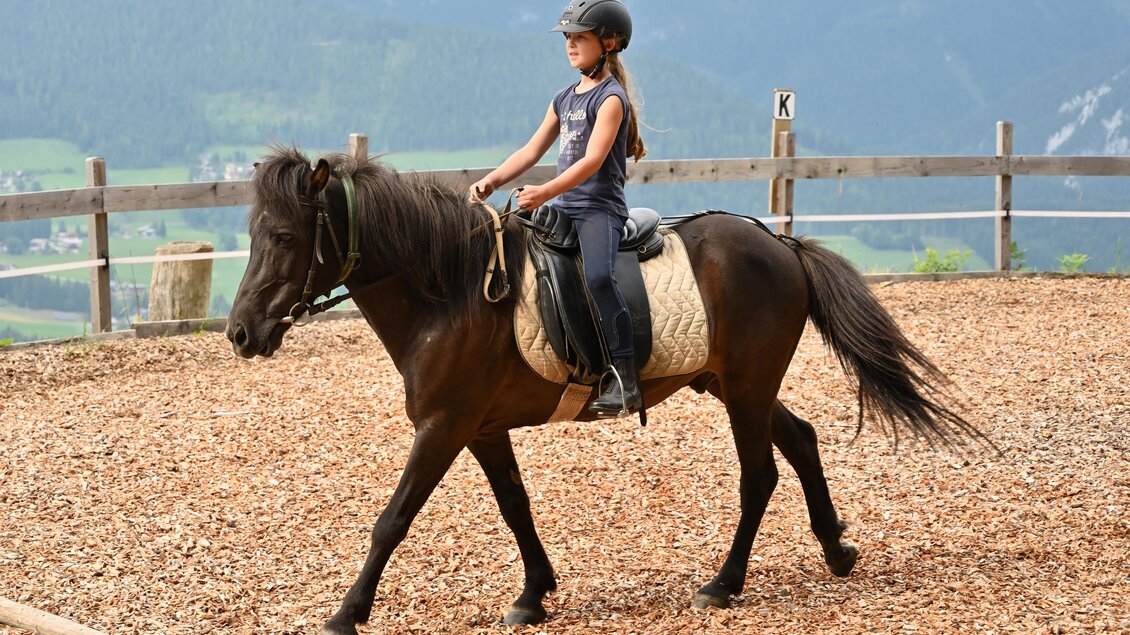 Ein Mädchen reitet auf einem schwarzen Pony in einer Reithalle. Im Hintergrund sind sanfte Hügel und Berge zu sehen. | © Breilerhof