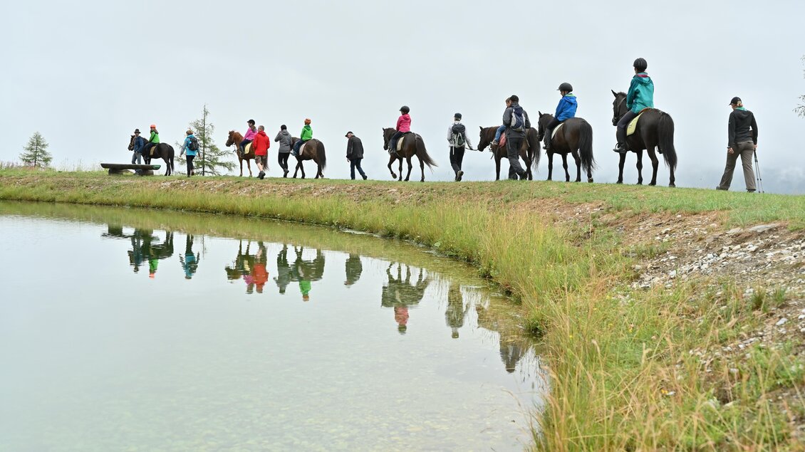 Eine Gruppe von Reitern bewegt sich entlang eines Teiches in einer bewölkten Landschaft. Die Reflexionen im Wasser sind deutlich sichtbar. | © Breilerhof