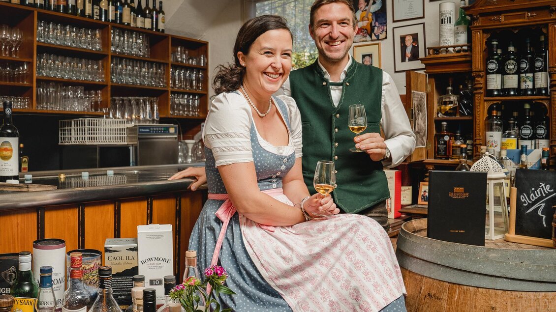 Ein fröhliches Paar steht an einer Bar. Die Frau trägt ein traditionelles bayerisches Dirndl und der Mann einen Trachtenanzug. | © www.dreihasen.at