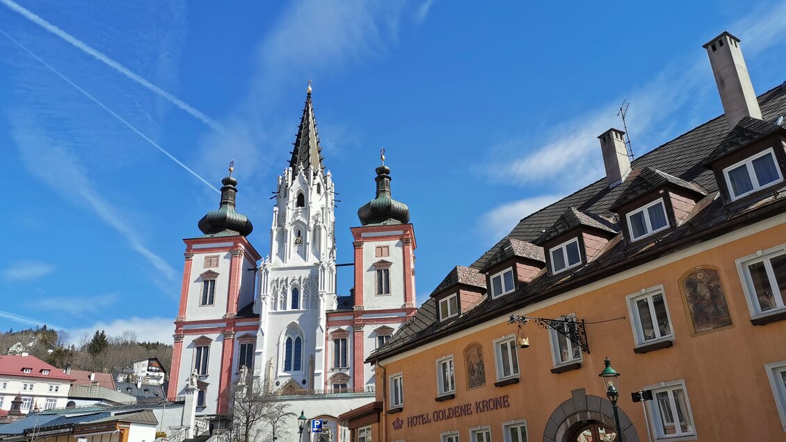 Eine beeindruckende Kirche mit einem hohen Turm und bunten Fassaden. Im Vordergrund sind einige Autos geparkt und der Himmel ist klar und blau. | © TV Hochsteiermark / Brigitte Digruber