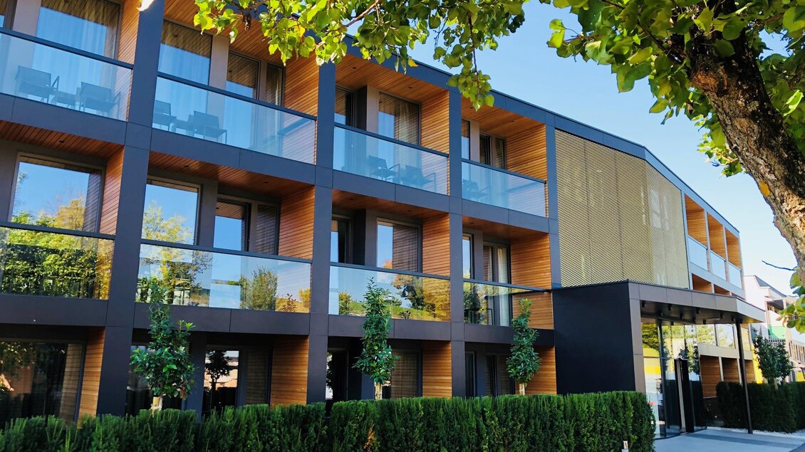 Ein modernes Gebäude mit mehreren Etagen und großen Fensterfronten. Umgeben von grünen Pflanzen und Bäumen, wirkt es einladend und freundlich. | © Thermenhotel Kowald