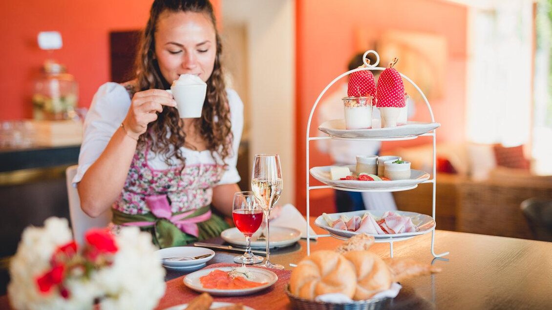 Eine Frau genießt eine Tasse Kaffee in einem gemütlichen Raum. Auf dem Tisch stehen köstliche Snacks und eine dekorative Etagere mit verschiedenen Leckereien.