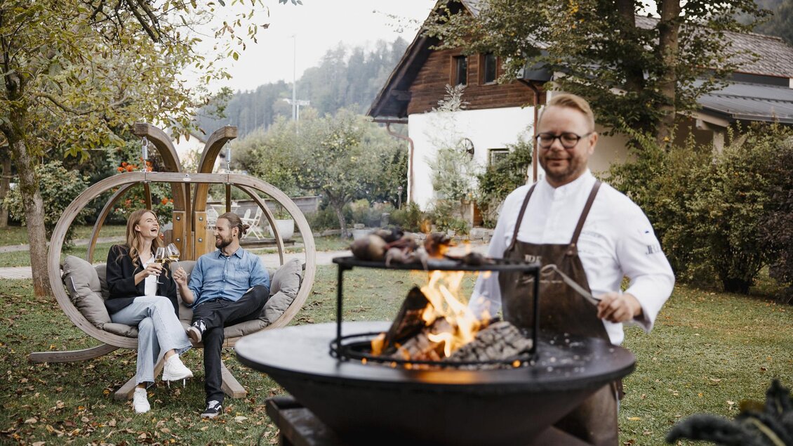 Ein Koch bereitet Grillgerichte über offenem Feuer vor. Im Hintergrund entspannen sich ein Paar in einer Hängematte. | © nicoleseiser.at