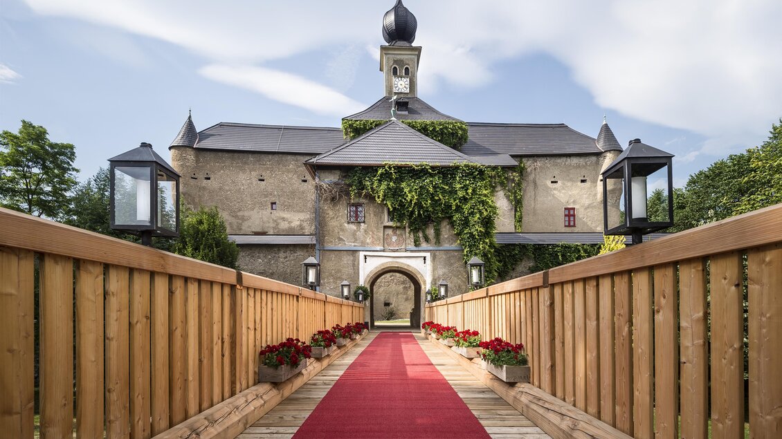 Ein historisches Schloss mit einer roten Teppichzufahrt. Die Fassade ist von grünem Efeu umrahmt und von Laternen gesäumt. | © Tauroa GmbH