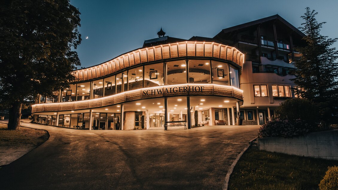 Ein modernes Gebäude mit beleuchteter Fassade und großen Fenstern. Der Schwagerhof ist von Bäumen umgeben und bei Dämmerung fotografiert. | © Hotel Schwaigerhof