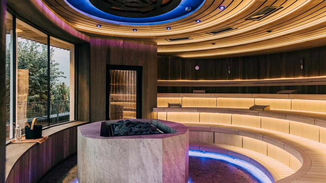 Ein moderner Wellnessbereich mit sanften Holzoberflächen und stimmungsvollem Licht. In der Mitte befindet sich ein runder Steintisch, umgeben von eleganten Sitzbereichen. | © Hotel Schwaigerhof
