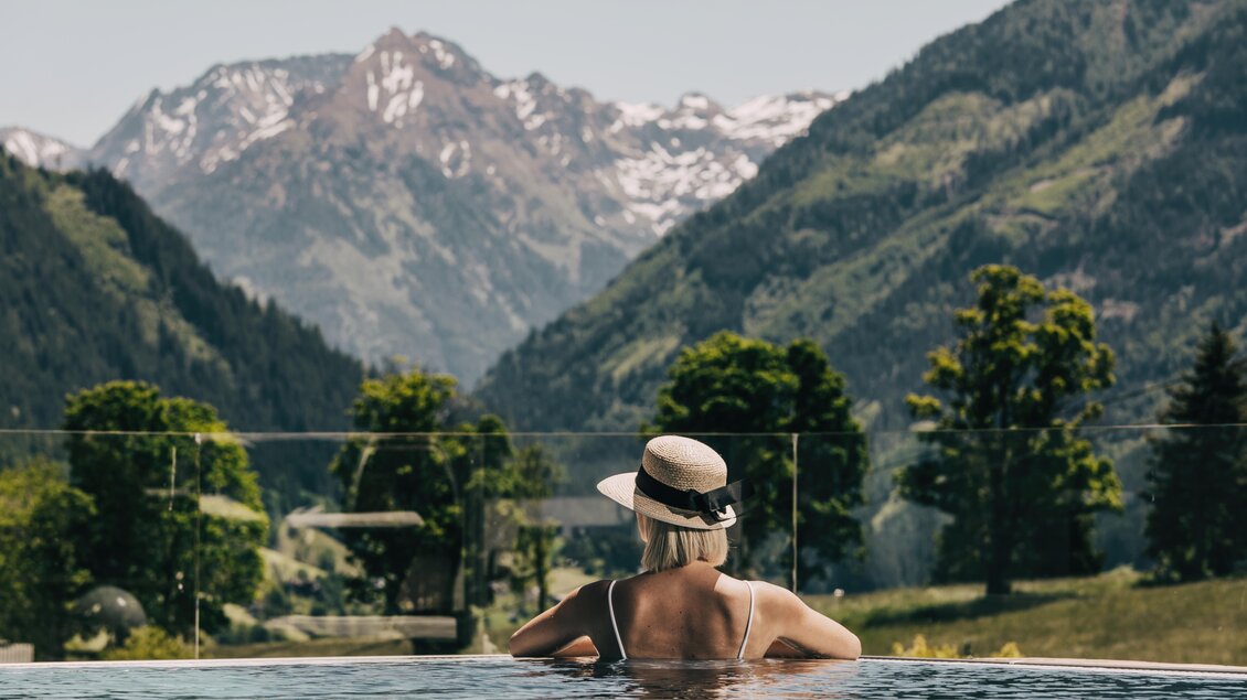 Eine Person sitzt entspannt im Pool und blickt auf die majestätischen Berge. Die Landschaft ist grün und von Bäumen umgeben, während der Himmel klar ist. | © Hotel Schwaigerhof