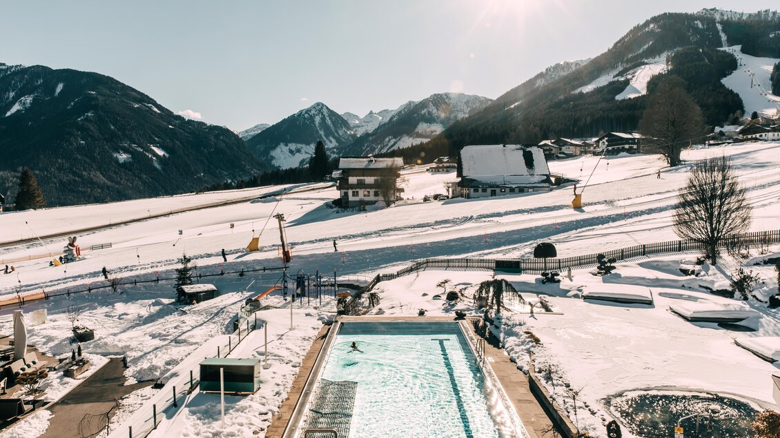Ein schneebedecktes Tal mit Bergblick und Skifahrern auf den Pisten. Im Vordergrund ist ein Schwimmbecken, das in die winterliche Landschaft integriert ist. | © Hotel Schwaigerhof