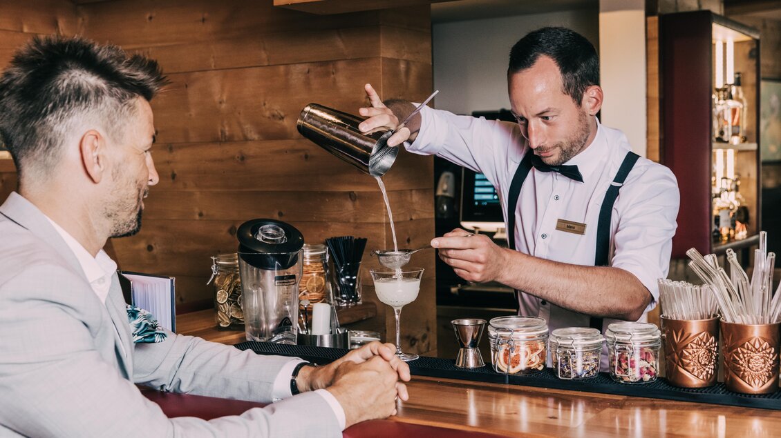 Ein Barkeeper, der einen Cocktail in ein Glas gießt, steht an der Bar. Ein Gast sitzt am Tisch und beobachtet aufmerksam. | © Hotel Schwaigerhof