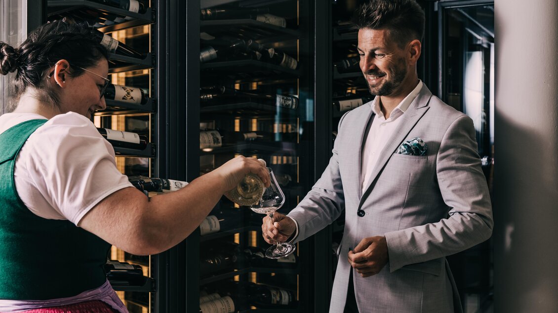 Eine Frau im Trachtenkleid schenkt einem Mann in einem Anzug ein Glas Wein ein. Im Hintergrund sind viele Weinflaschen in einem Kühlschrank zu sehen. | © Hotel Schwaigerhof