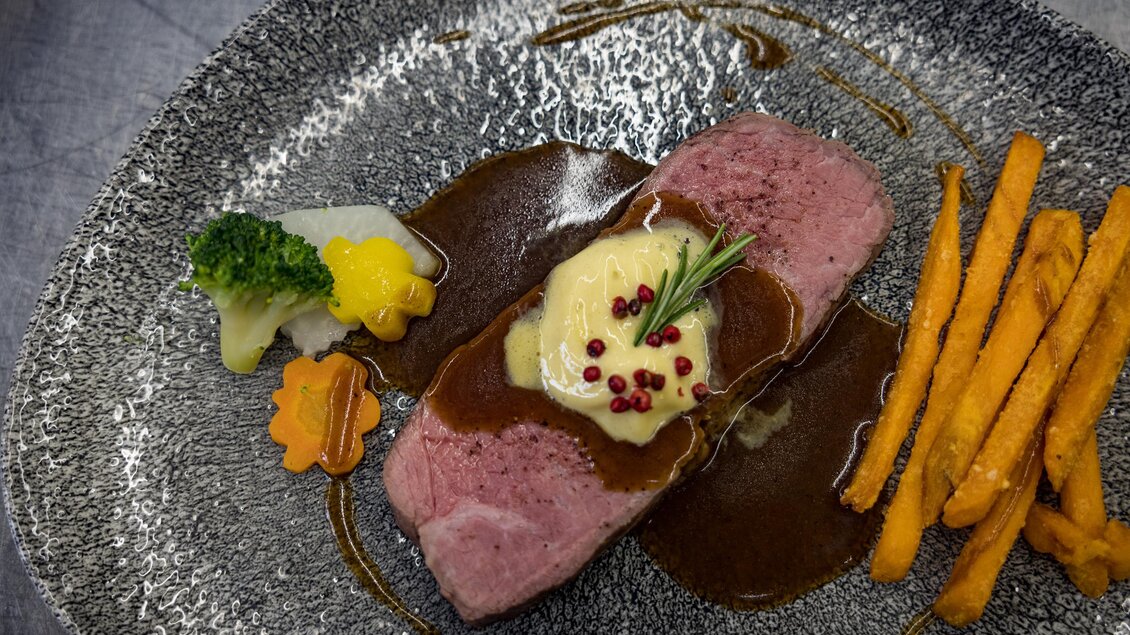 Ein ansprechend angerichtetes Stück Fleisch mit einer dunklen Soße, umgeben von buntem Gemüse und Kartoffelstäbchen. Das Gericht ist elegant auf einem dekorativen Teller präsentiert. | © Netzwerk Kulinarik wildbild