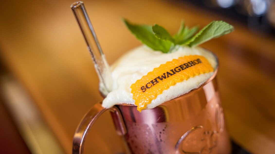 Ein Getränk in einem kupfernen Becher mit einer Schicht Sahne, einem Strohhalm und einer limettengrünen Minze. Auf der Getränkedekoration befindet sich ein orangefarbenes Band mit dem Schriftzug "SCHWAIERGEN." | © Netzwerk Kulinarik wildbild