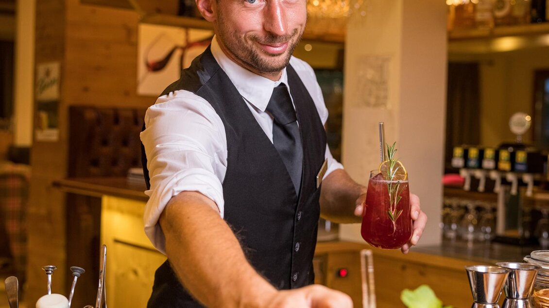 Ein Barkeeper serviert einen Drink an der Bar. Er lächelt freundlich und trägt eine elegante Weste. | © Netzwerk Kulinarik wildbild