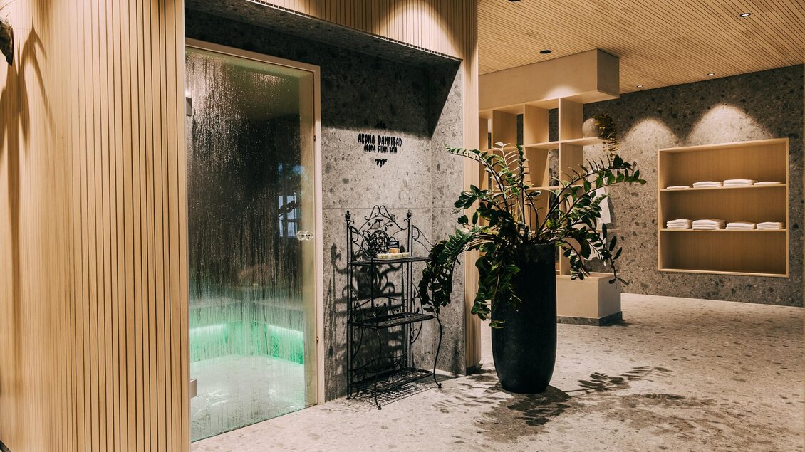 Ein moderner Wellnessbereich mit einer Dampfsauna und einem großen, grünen Pflanzentopf. Die Wände sind in Holzoptik gehalten und es gibt Regale mit Handtüchern im Hintergrund. | © Hotel Schwaigerhof