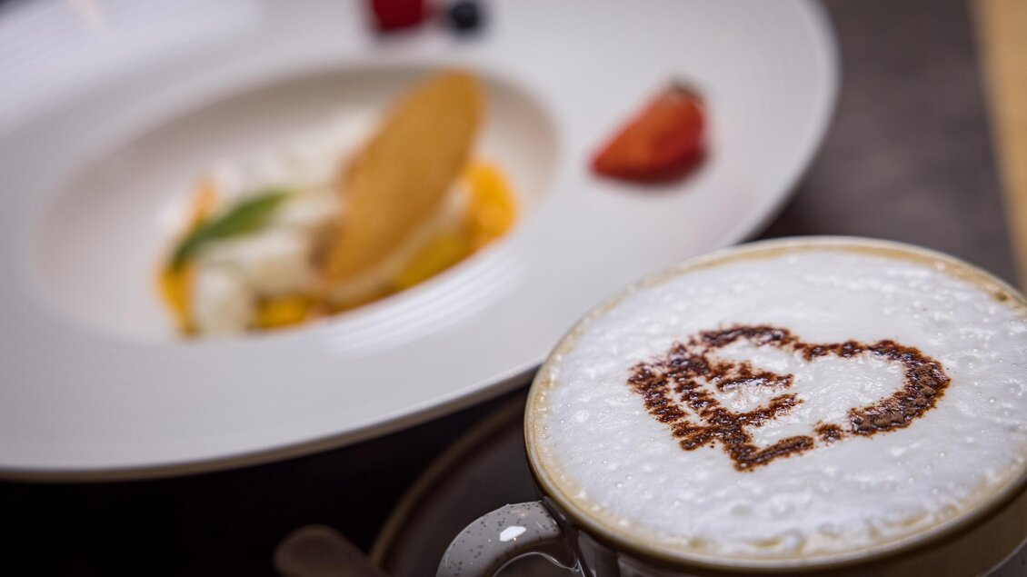 Eine Tasse Café mit einem herzförmigen Muster aus Schokolade auf dem Schaum. Im Hintergrund sieht man ein Dessert mit Früchten und einer Keksspezialität. | © Netzwerk Kulinarik wildbild
