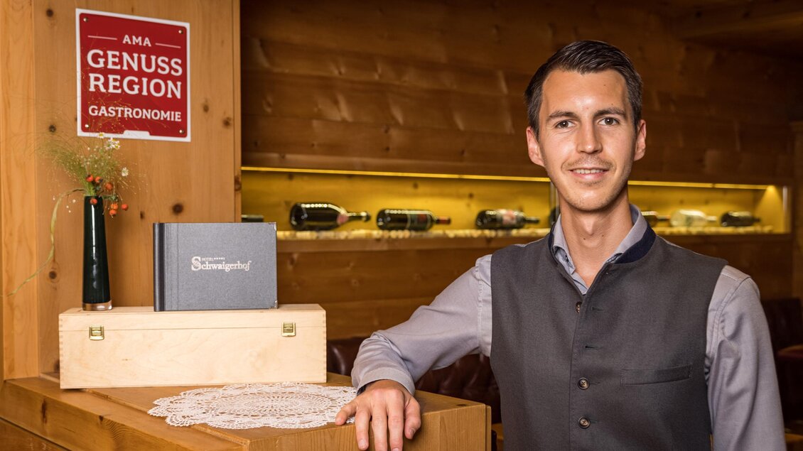 Ein lächelnder Mann in traditioneller Tracht steht in einem Holzraum. Im Hintergrund sind Weine und eine Tafel mit dem Zeichen „Genussregion Gastronomie“ sichtbar. | © Netzwerk Kulinarik wildbild