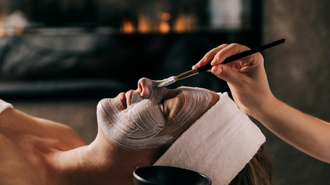 Eine Person erhält eine Gesichtsmaske in einem entspannenden Umfeld. Im Hintergrund ist ein warmes Licht zu sehen, das eine gemütliche Atmosphäre schafft. | © Hotel Schwaigerhof