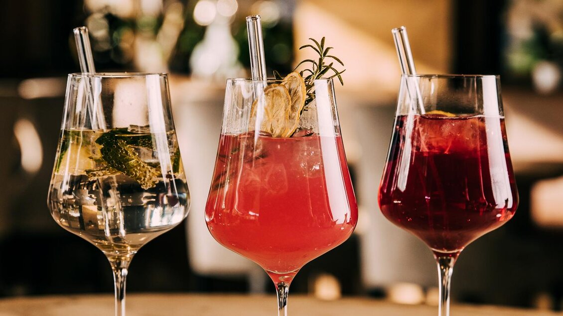 Drei elegante Cocktails auf einem Holztisch. Der linke Cocktail ist klar und enthält frische Kräuter, während die anderen zwei pink und rot sind, mit verschiedenen Garnierungen. | © Hotel Schwaigerhof