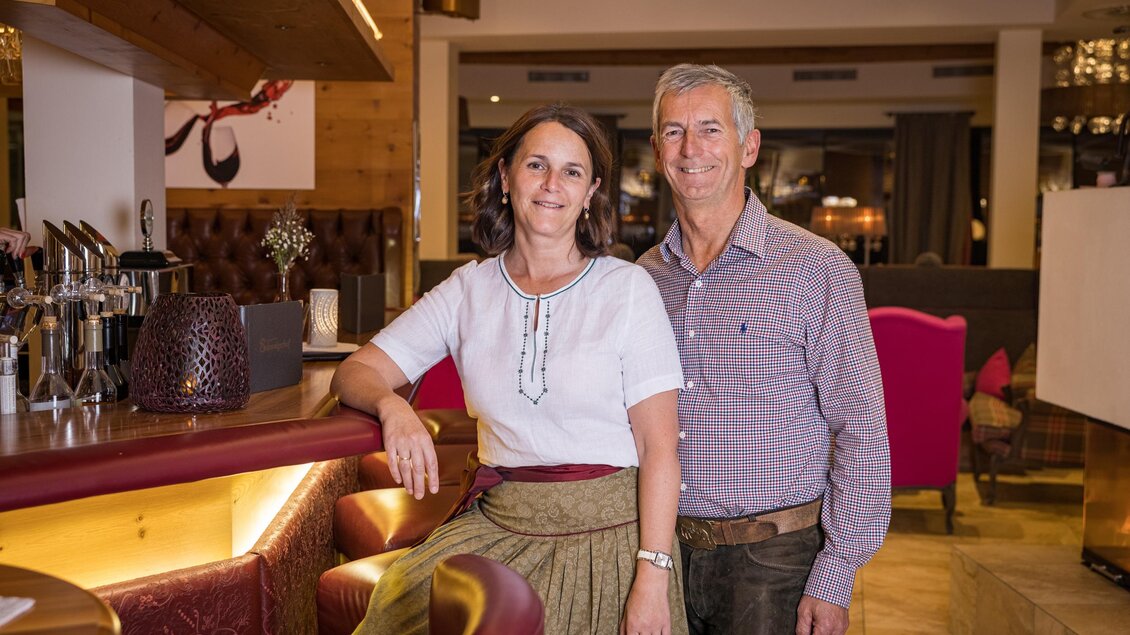 Ein freundliches Paar steht an der Bar in einem gemütlichen Restaurant. Die Frau trägt ein traditionelles Kleid und der Mann lächelt in einem Hemd. | © Netzwerk Kulinarik wildbild