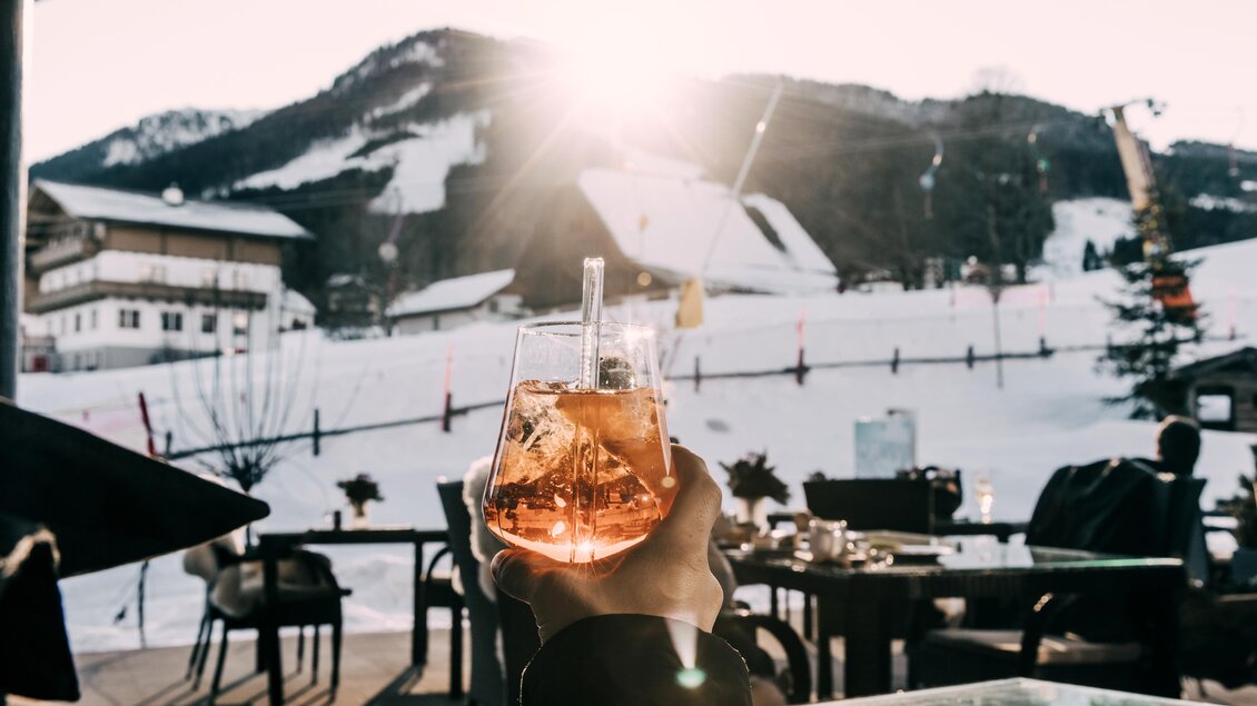 Ein Hand hält ein Glas mit einem orangefarbenen Getränk vor einer winterlichen Berglandschaft. Die Sonne scheint und es sind Skilifte im Hintergrund sichtbar. | © Hotel Schwaigerhof