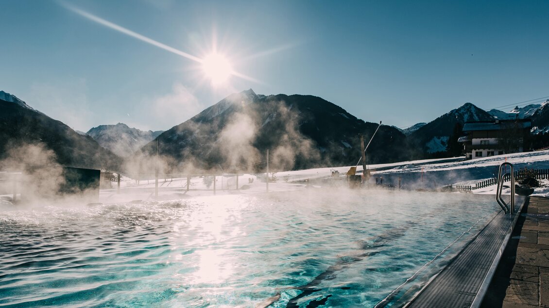 Ein beheizter Außenpool in den Bergen, umgeben von schneebedeckten Gipfeln. Die Sonne strahlt am blauen Himmel und Dampf steigt über dem Wasser auf. | © Hotel Schwaigerhof