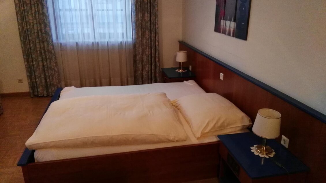 Ein gemütliches Schlafzimmer mit einem Doppelbett und Nachttischen. Es gibt ein Fenster mit Vorhängen und einen warmen Holzfußboden. | © Hotel Seltenriegel